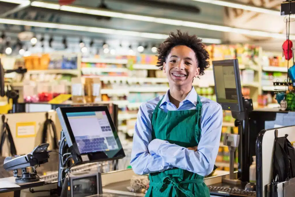 A young grocery store cashier standing with his arms folded smiling at the camera